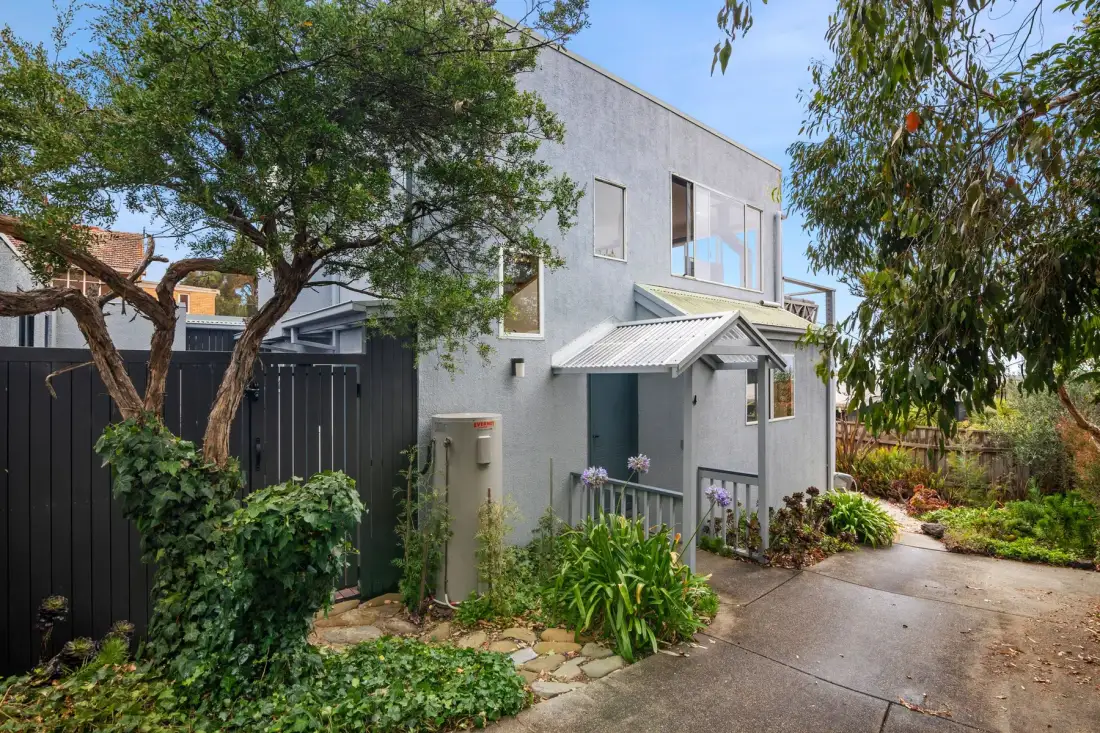 A modern two-story house stands beside a paved path and landscaped garden.