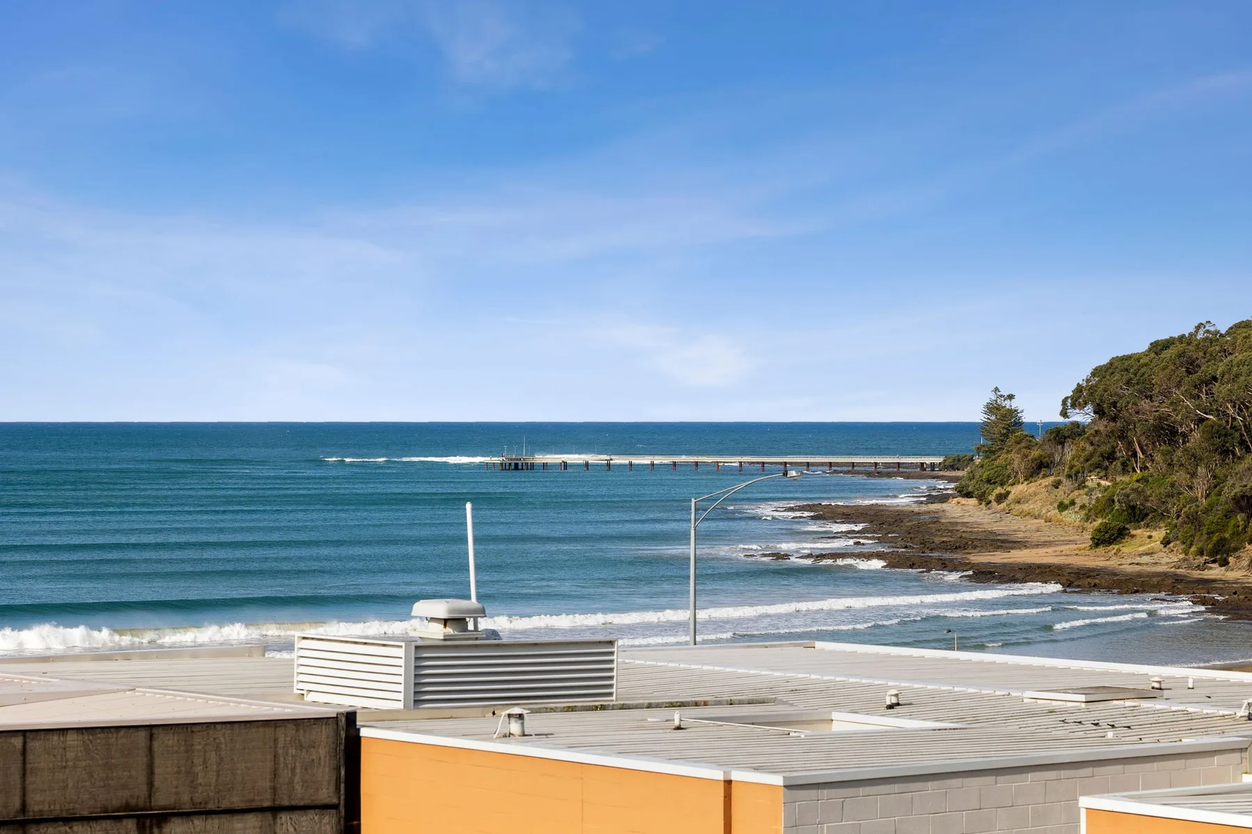 Coastal seascape with ocean waves, a rocky shoreline, and nearby rooftops.