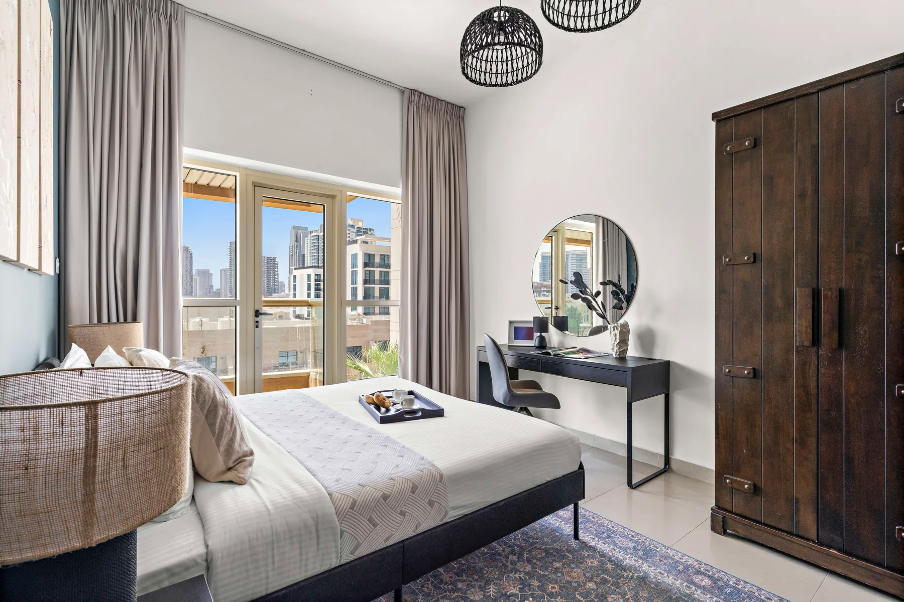 A bright bedroom with a bed, desk, and large windows overlooking nearby buildings.