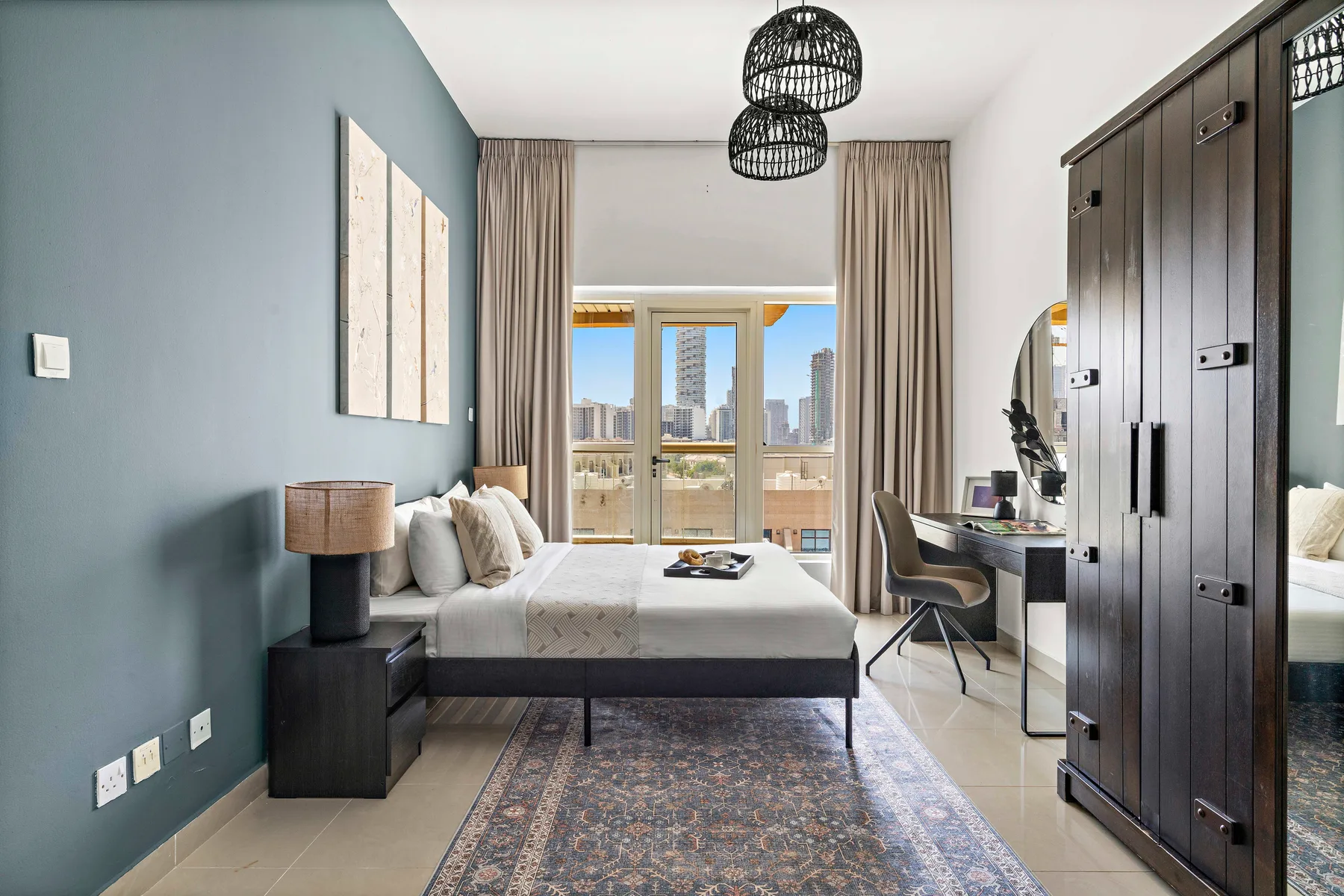 A bedroom with a bed, desk, and large windows overlooking city buildings.