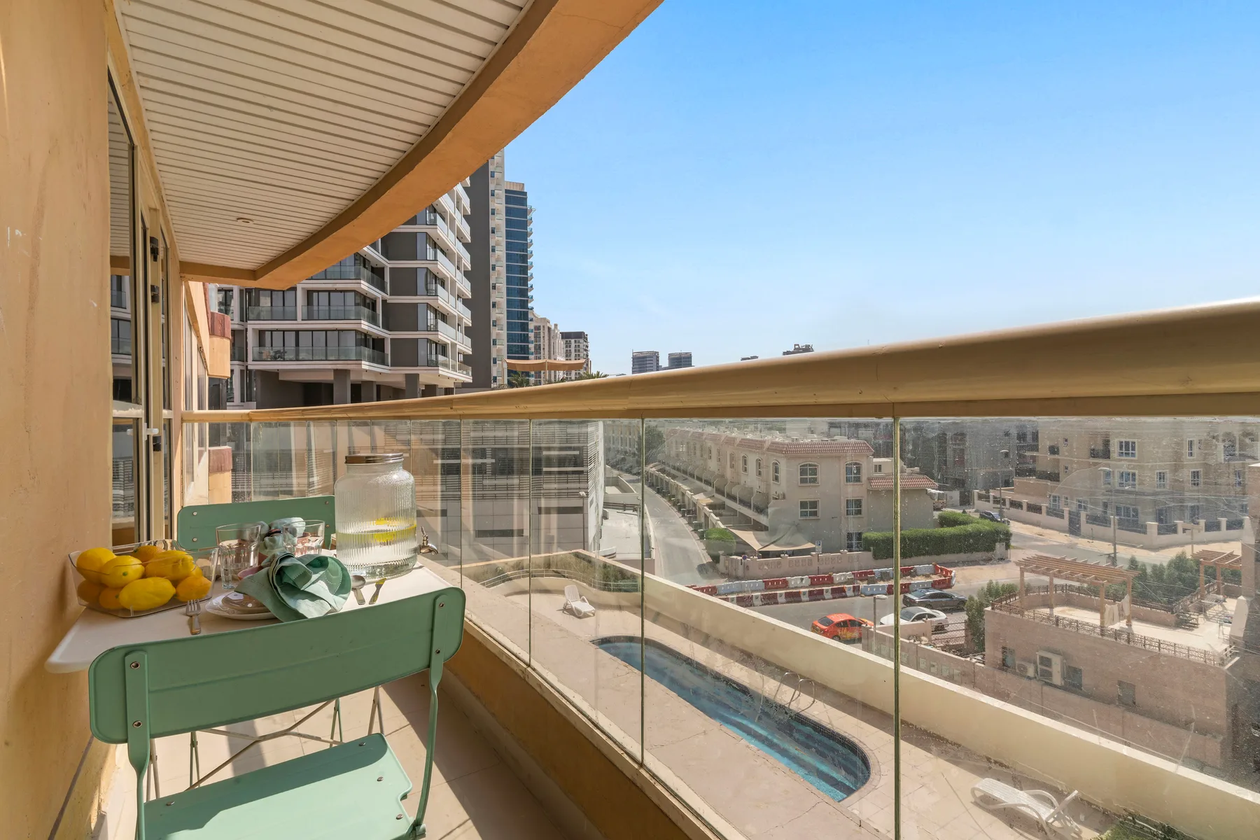 A balcony with a small table and seating overlooks nearby buildings and a pool.
