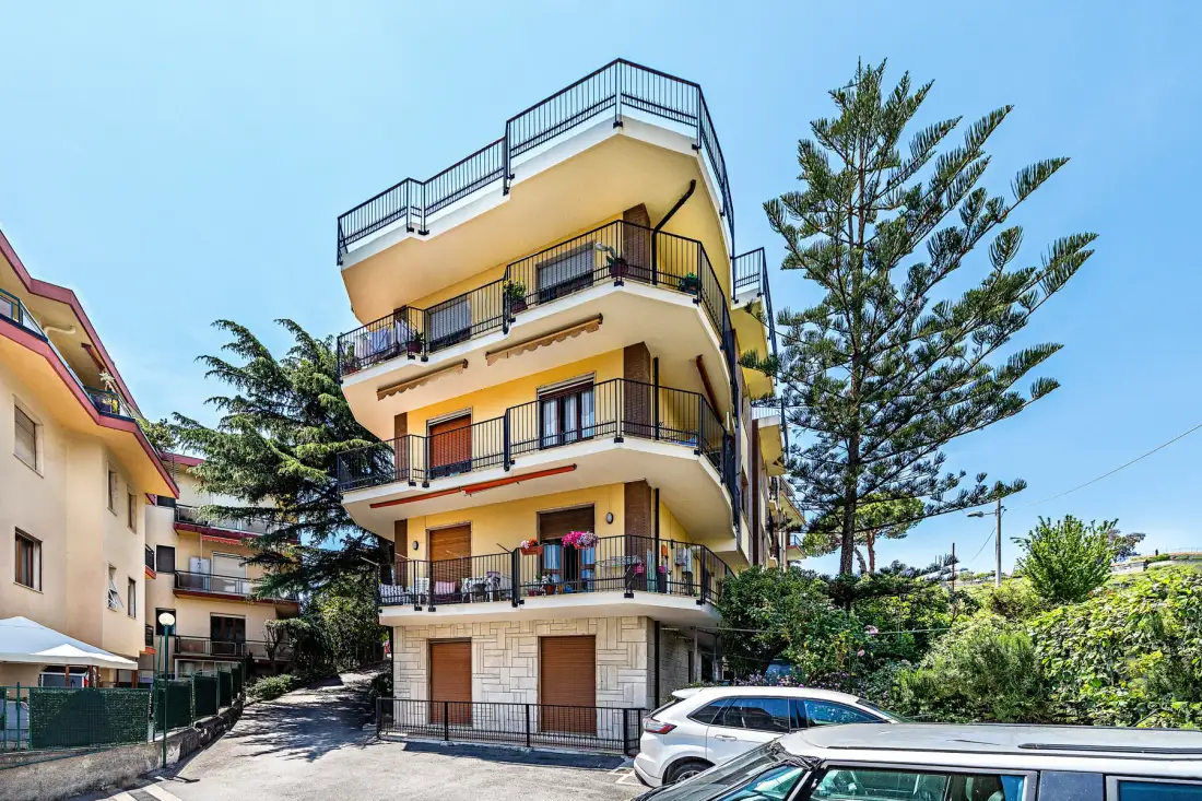 Un edificio de varias plantas con balcones está junto a una entrada y coches estacionados.