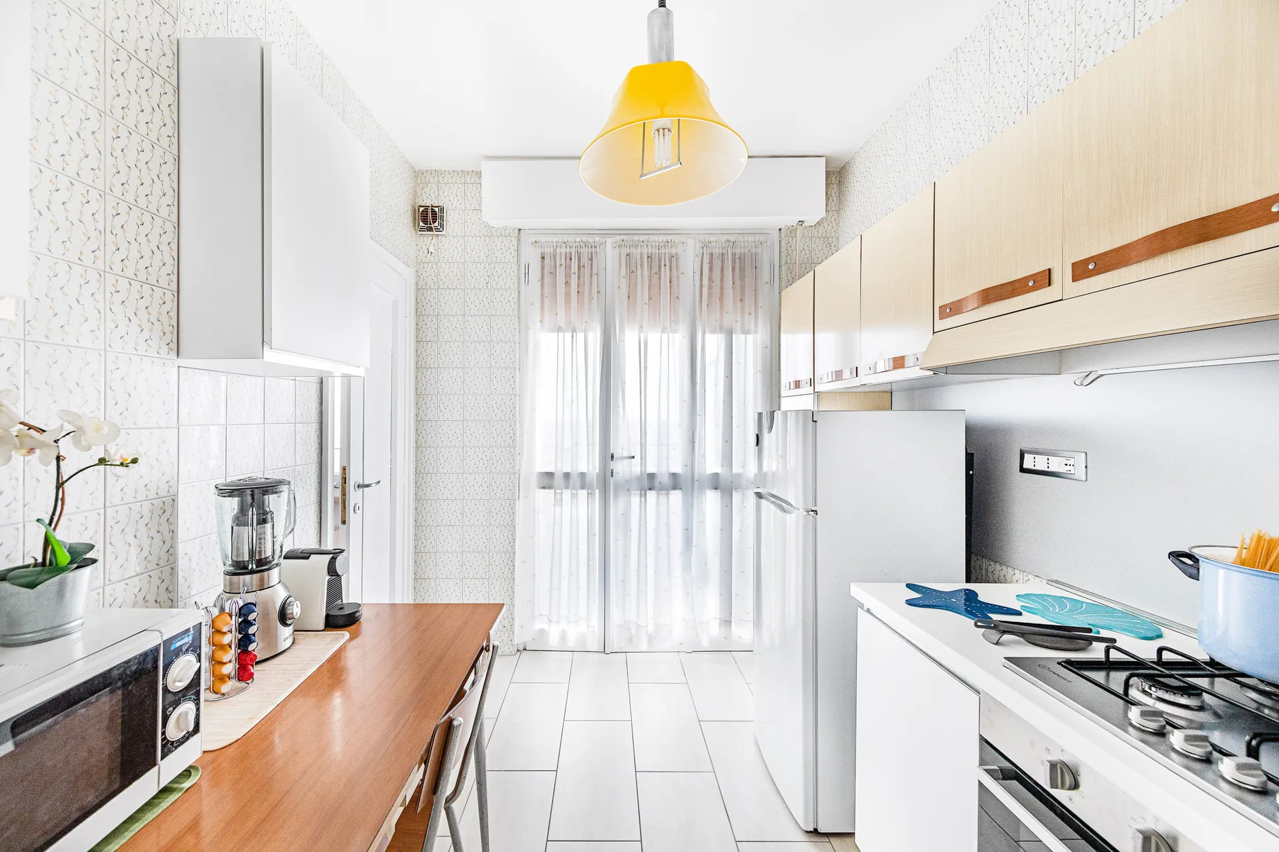 Una cocina luminosa con gabinetes claros, un refrigerador y una ventana con cortinas transparentes.