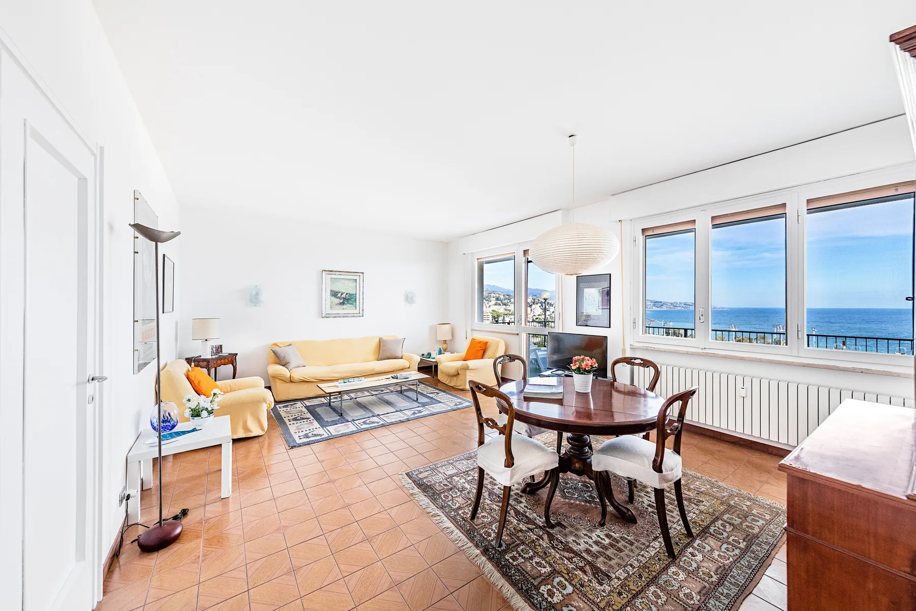 Un luminoso salón-comedor tiene asientos, una mesa de comedor y grandes ventanas con vistas al mar.
