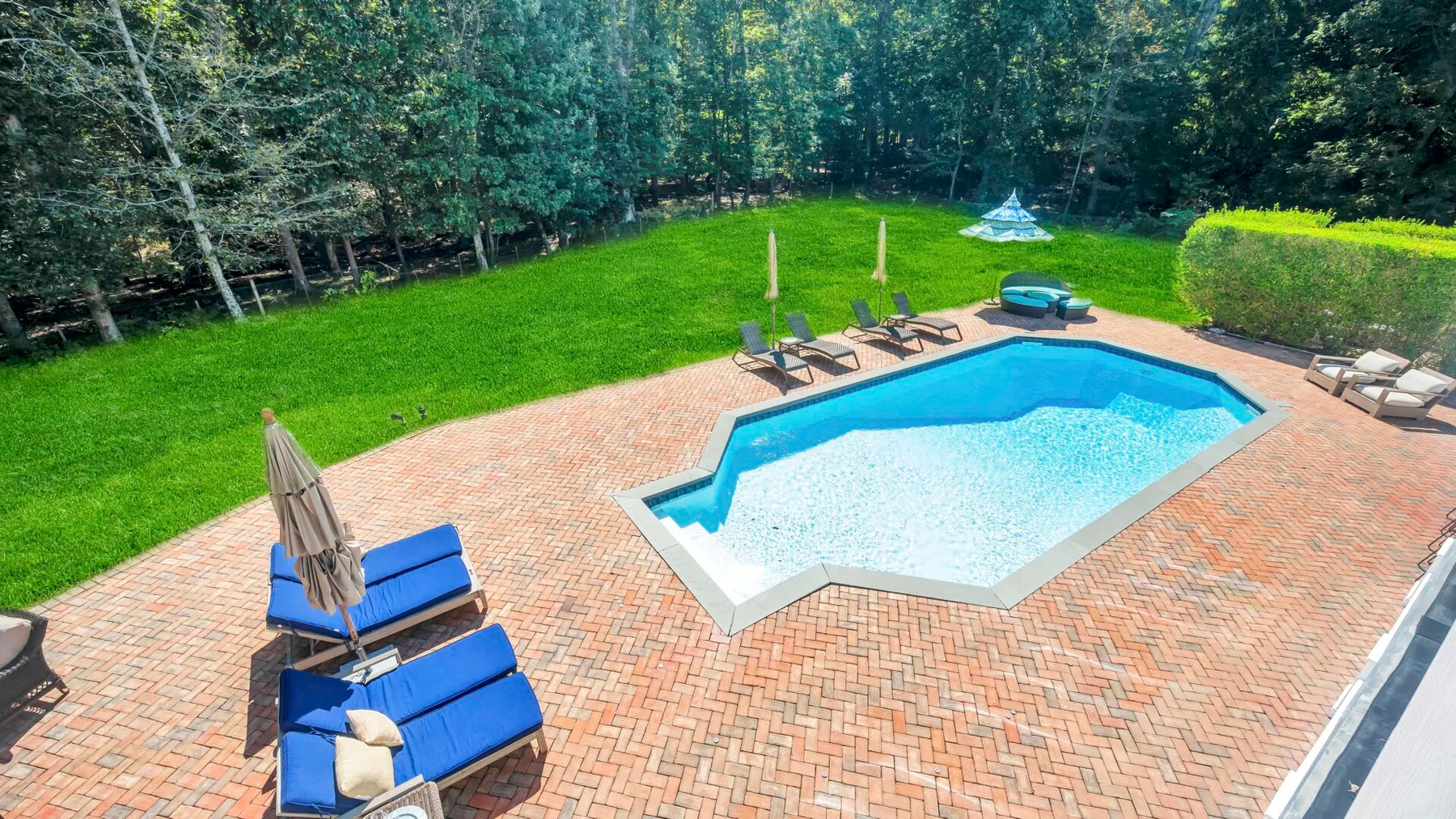 Una piscina al aire libre está en un patio de ladrillo, rodeada de césped y árboles.