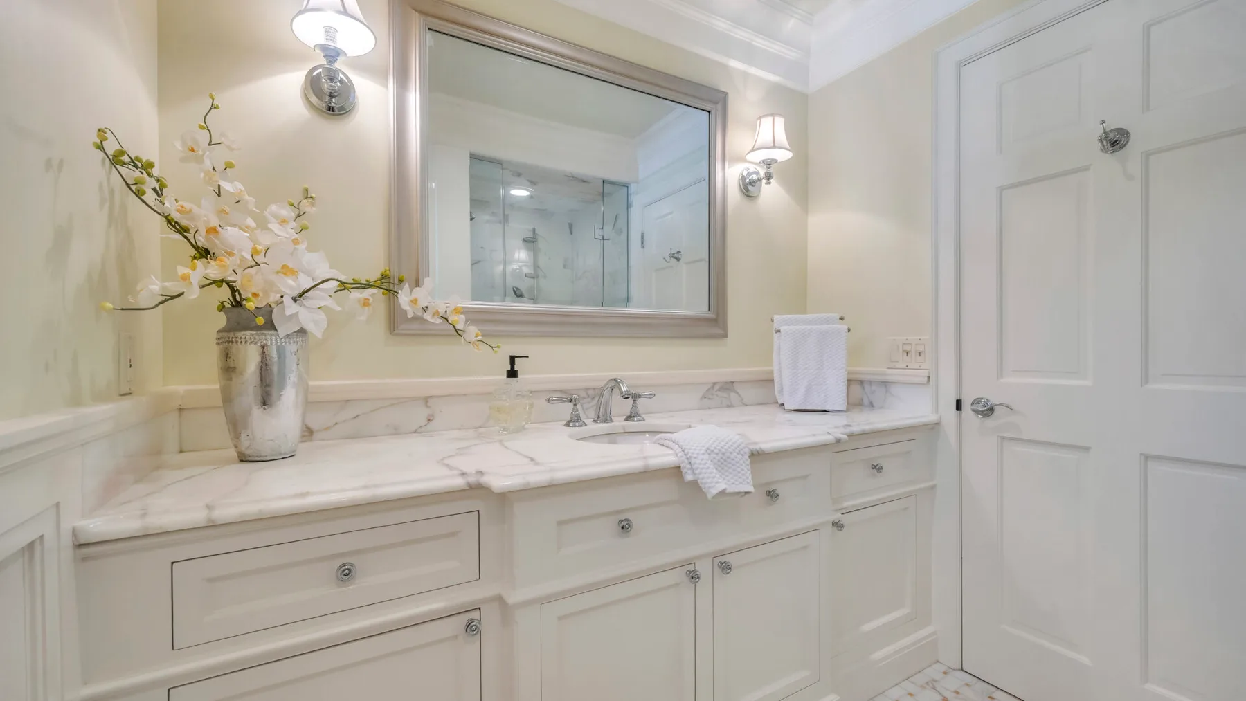 Un lavabo de baño luminoso con un espejo amplio y flores decorativas.