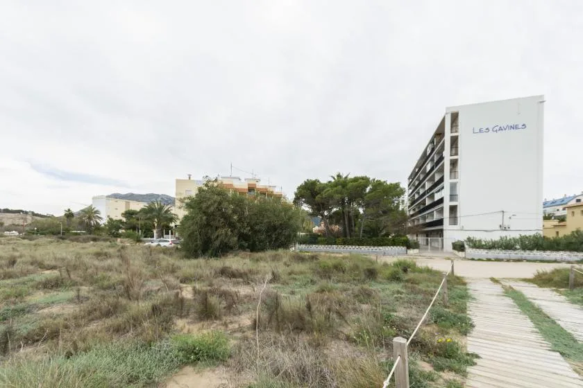 An apartment building stands beside a scrub-covered open lot and a wooden walkway.