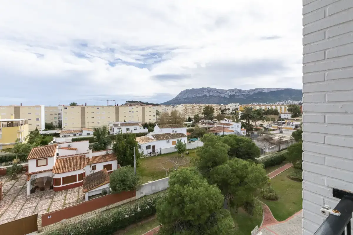 A balcony overlooks residential buildings, greenery, and distant mountains beneath a cloudy sky.