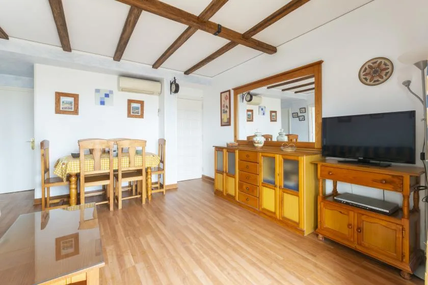 A bright living and dining area has wooden seating, a dining table, and a television.