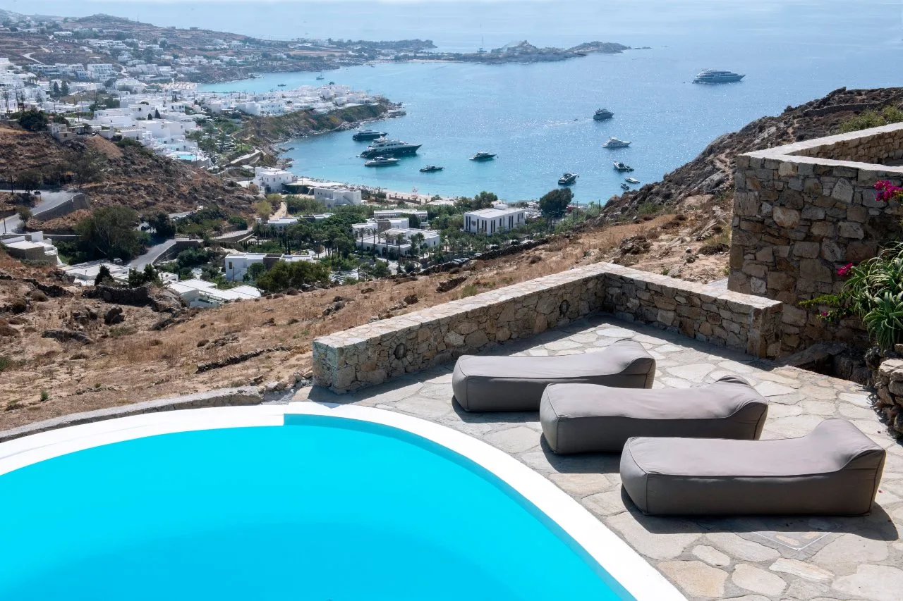 A terrace with a curved pool and loungers overlooks a coastal bay.