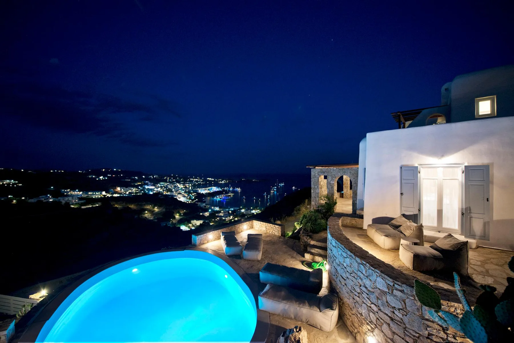 A lit swimming pool and terrace overlook a coastal town at night.