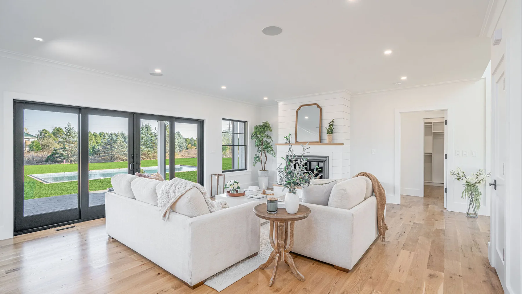 A bright living room with pale seating beside large windows overlooking a green yard.