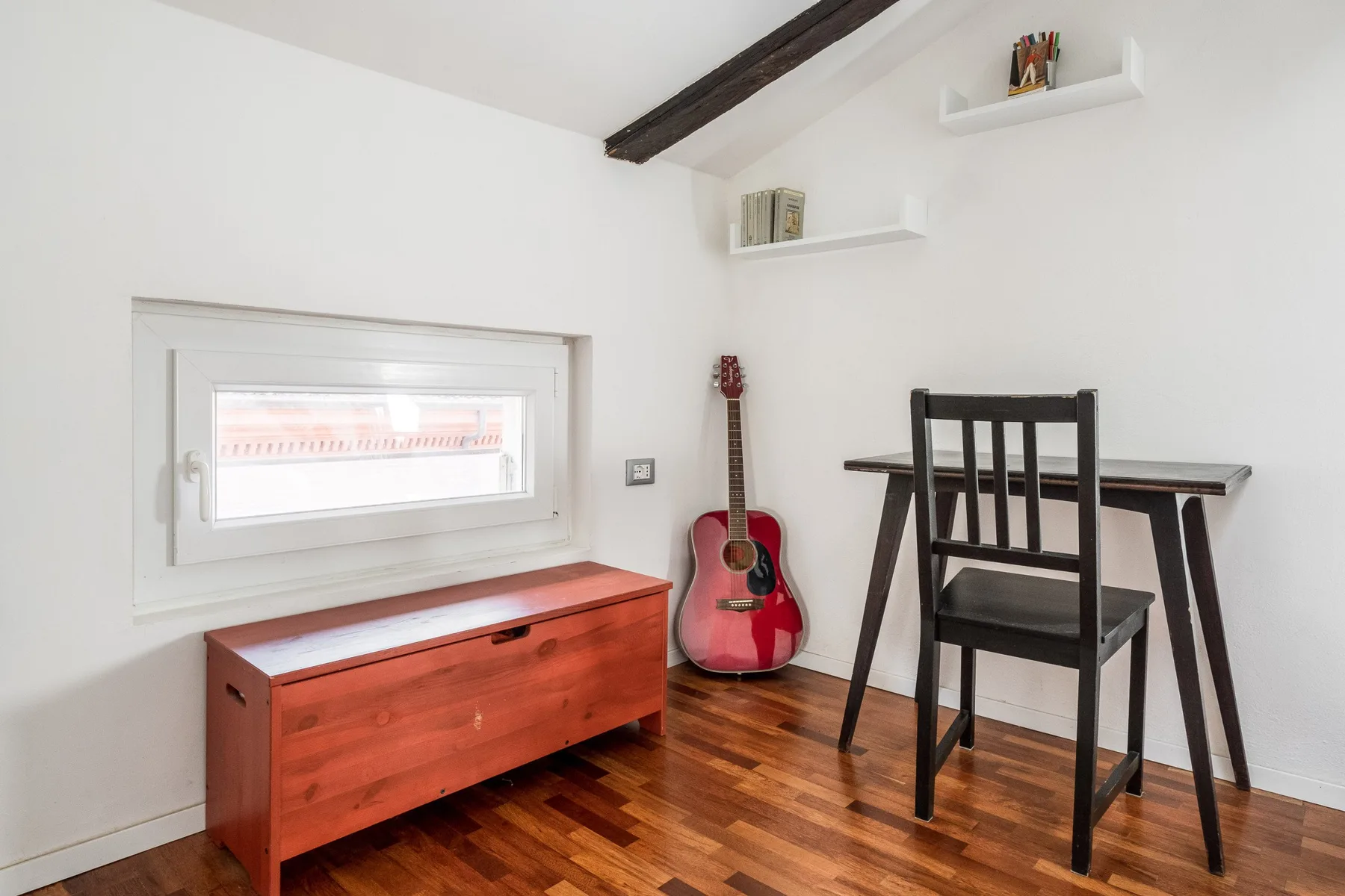 Una habitación luminosa con escritorio de madera, silla, guitarra y baúl bajo.