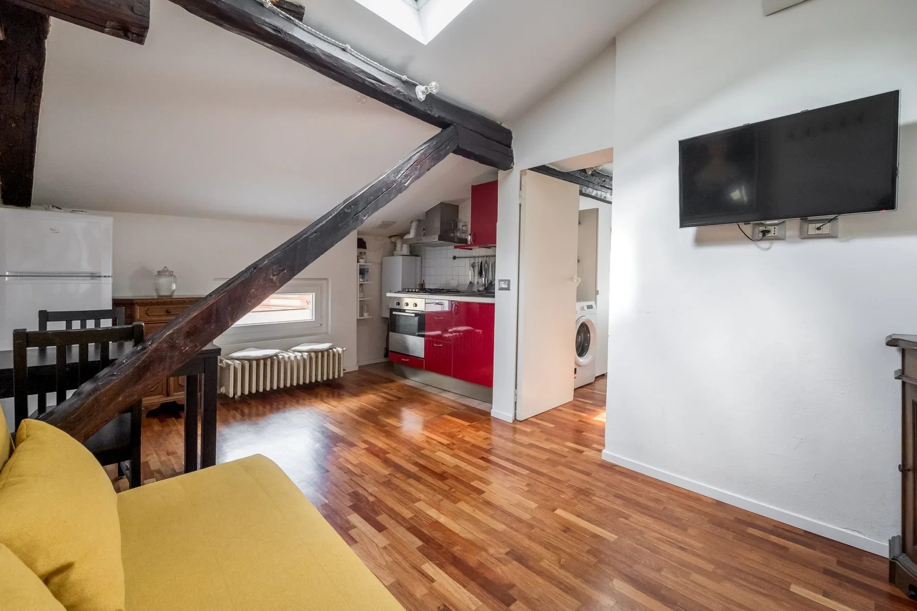 Una sala tipo loft con un sofá amarillo, vigas expuestas y una cocina compacta.