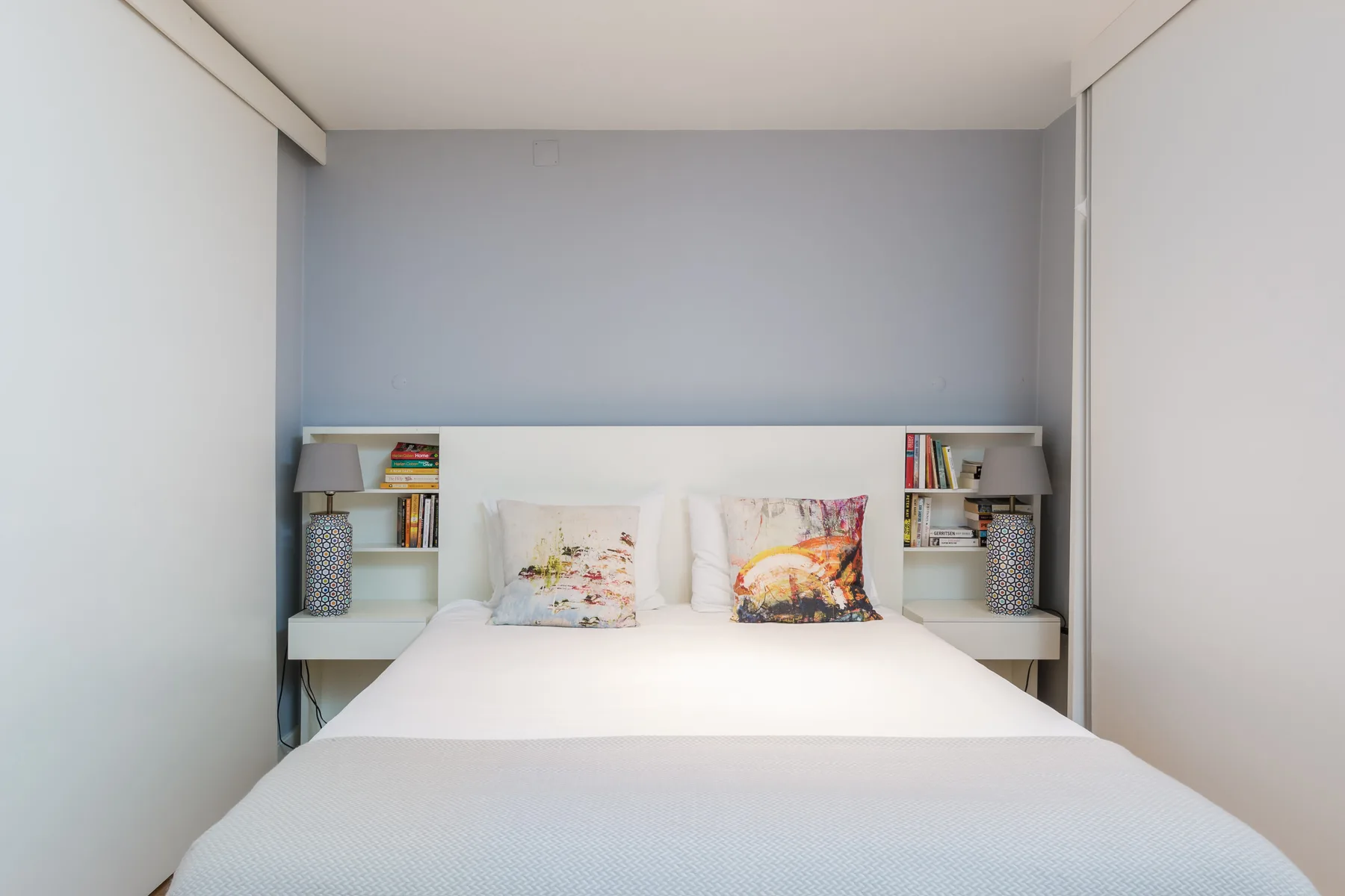 Un dormitorio con una cama blanca, cojines estampados y estantes empotrados.
