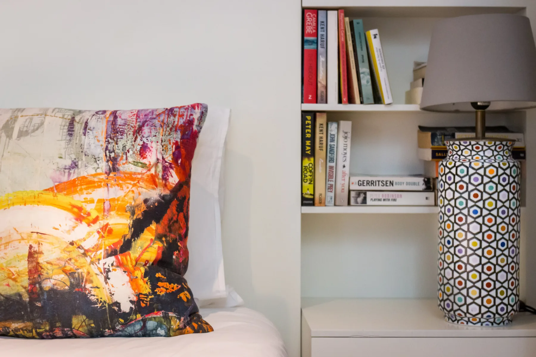 Un dormitorio con un cojín colorido junto a una estantería y una lámpara de mesa.