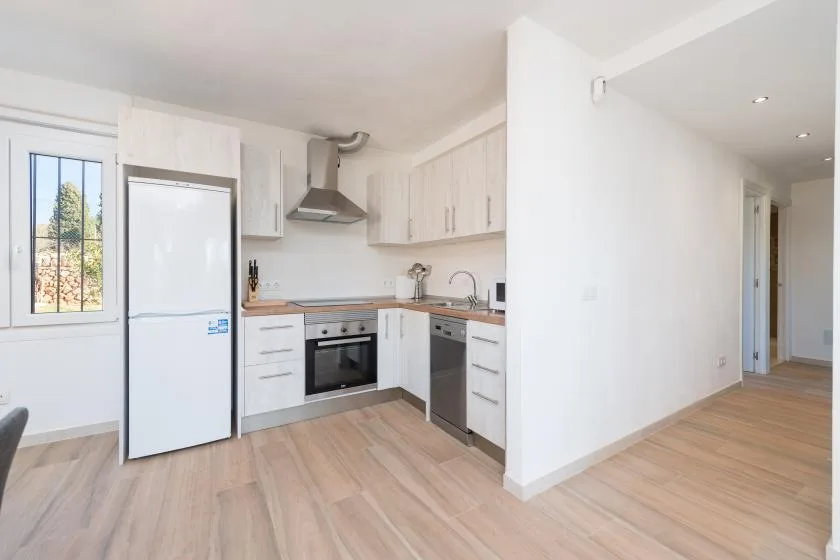 Una cocina luminosa con gabinetes blancos, electrodomésticos y piso de madera clara.