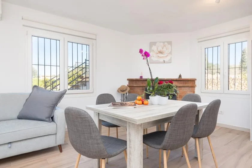 Un comedor luminoso con una mesa y asientos tapizados de color gris junto a grandes ventanas.