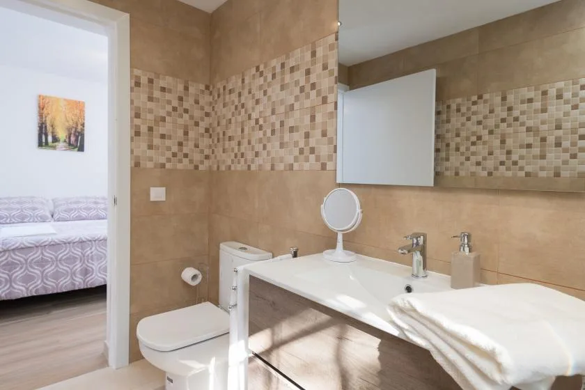 Un baño alicatado con lavabo, inodoro y una puerta abierta hacia un dormitorio.