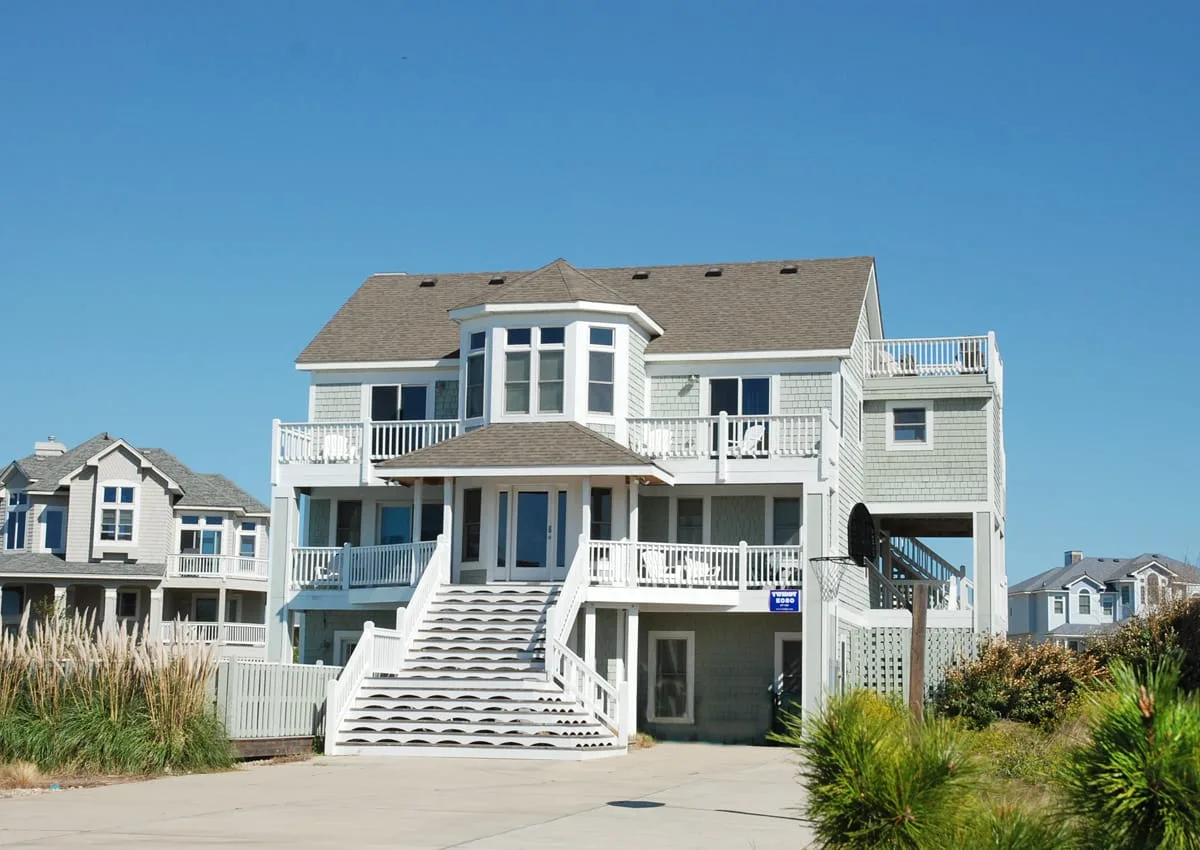 A large coastal-style house with balconies and a broad staircase faces the street.