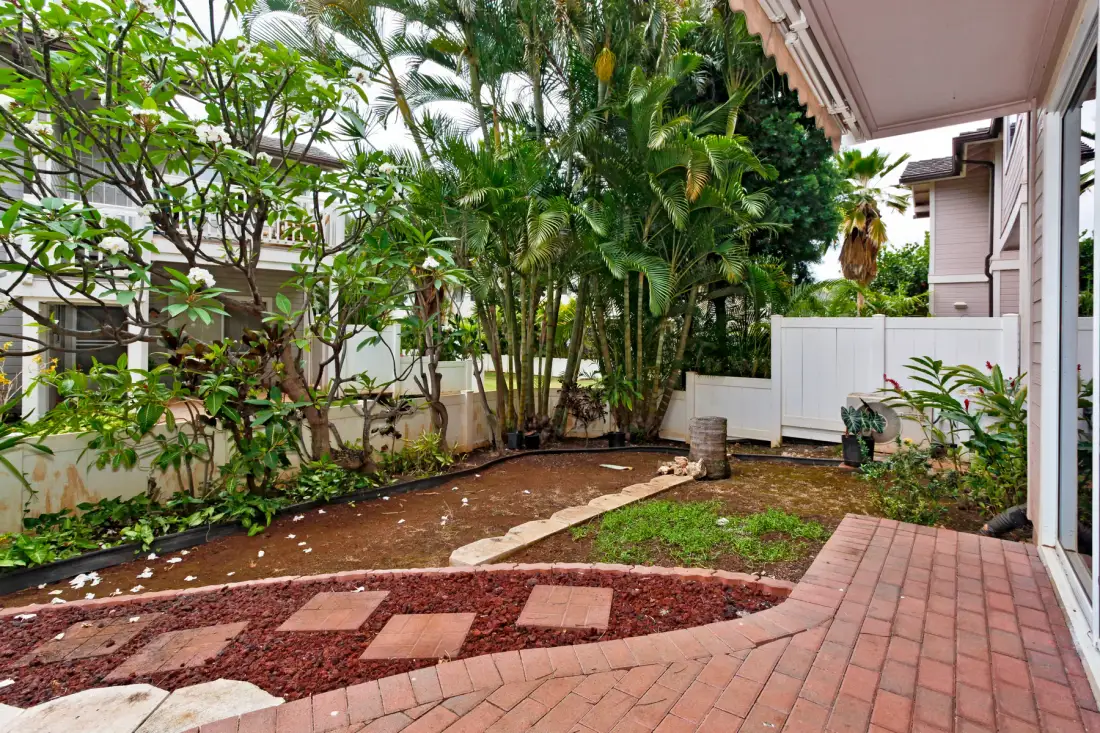 A landscaped garden beside a house, with tropical plants and a brick walkway.