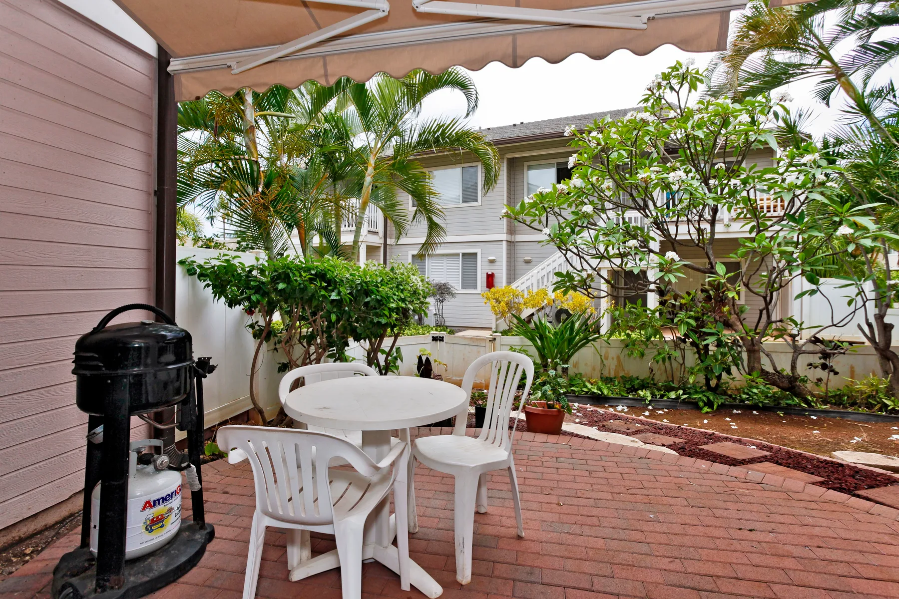 A brick patio with white outdoor seating sits beneath a canopy beside leafy plants.