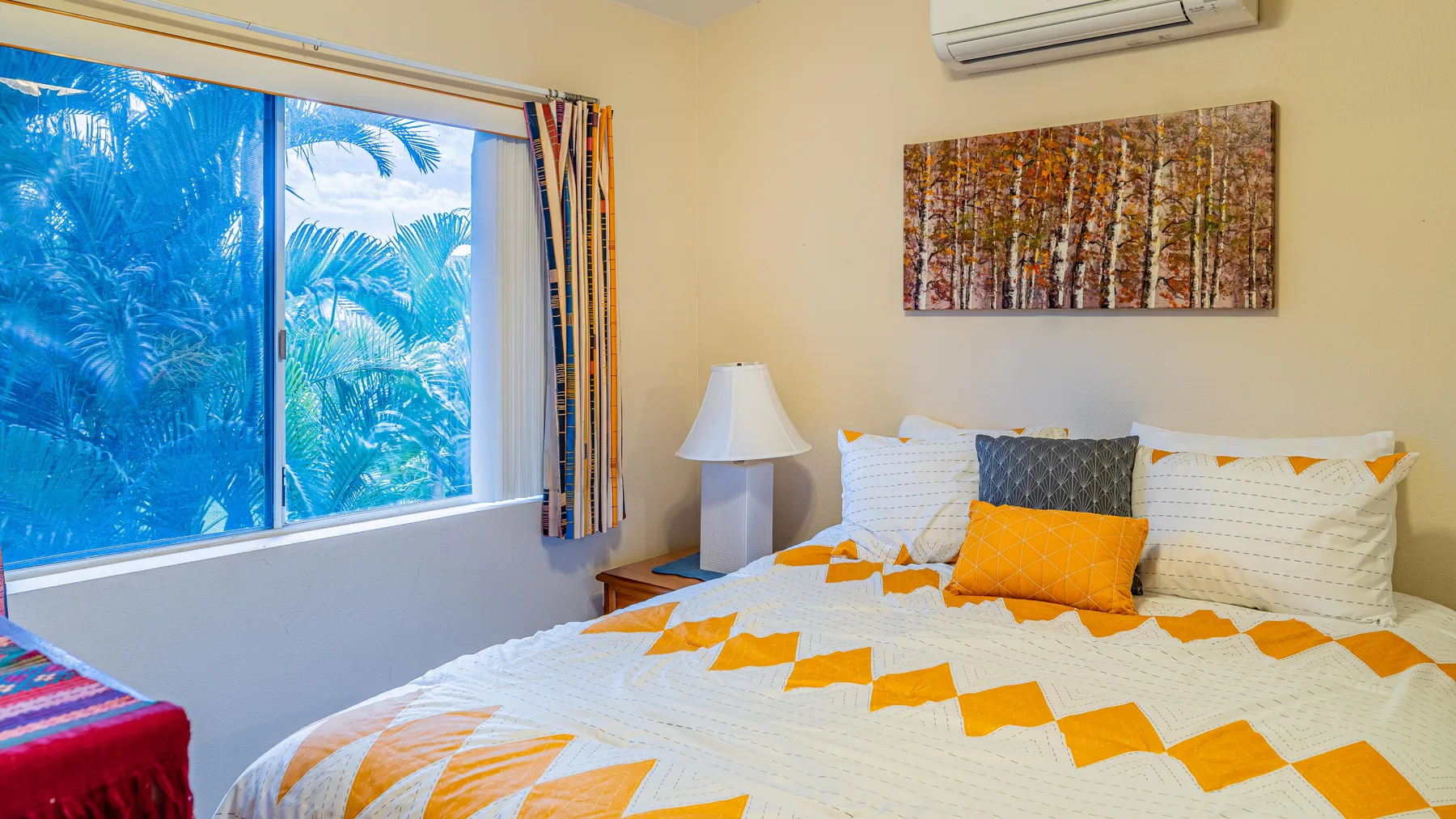 A bedroom with a bed dressed in white and orange bedding beside a large window.