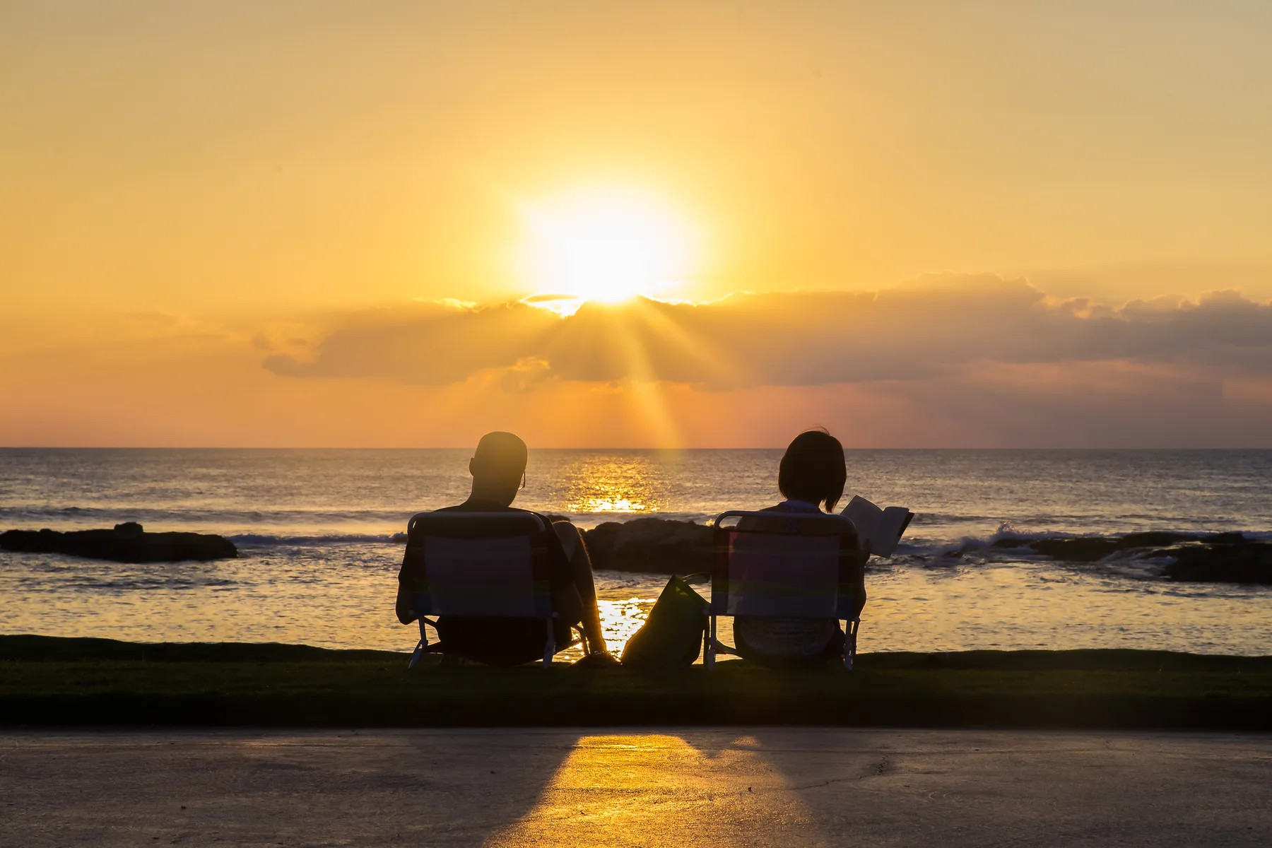 Two people sit by the ocean as the sun sets over the water.