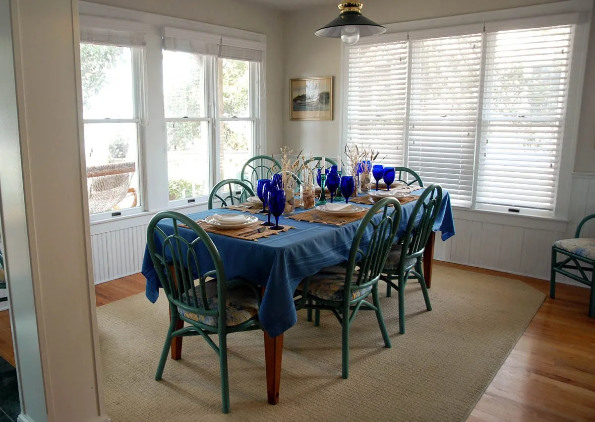 Un comedor tiene una mesa preparada con mantelería azul junto a grandes ventanas.
