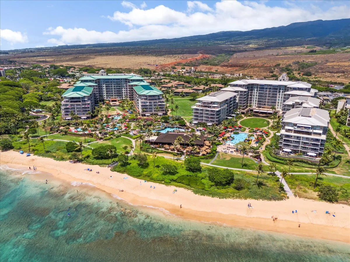 Vista aérea de un complejo junto a la playa, con jardines, piscinas y colinas alrededor.