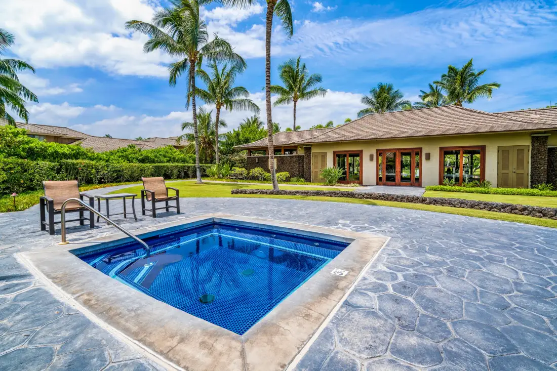 An outdoor swimming pool sits beside a paved patio, with a low building and palm trees behind it.