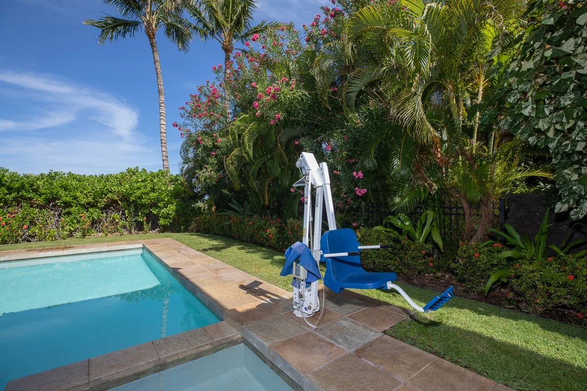 An outdoor swimming pool beside a blue pool lift, tropical plants, and a paved deck.