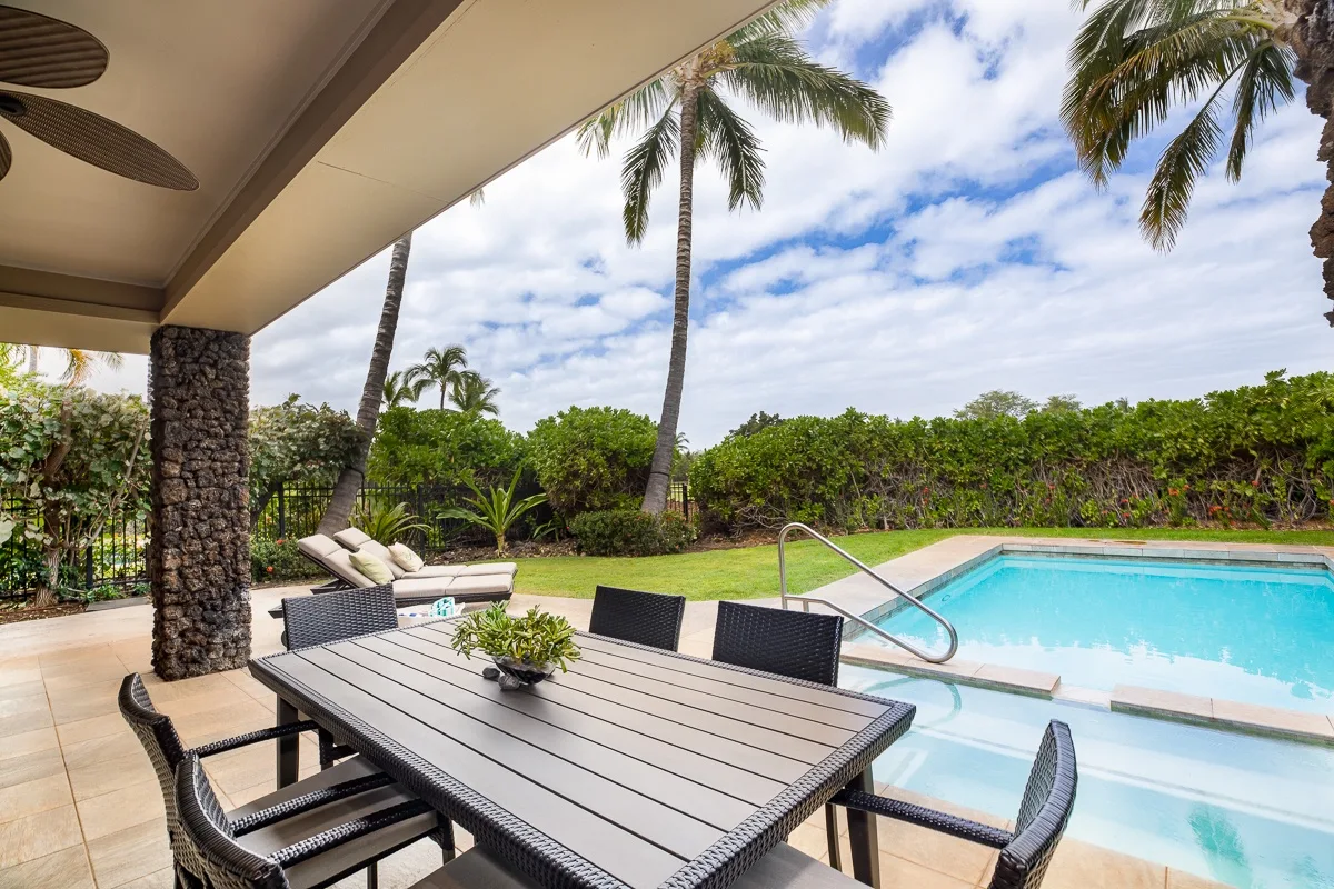 A patio dining area overlooks a swimming pool, with palm trees and greenery beyond.