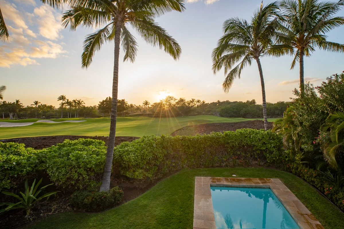 A swimming pool overlooks a green golf course beneath palm trees at sunset.