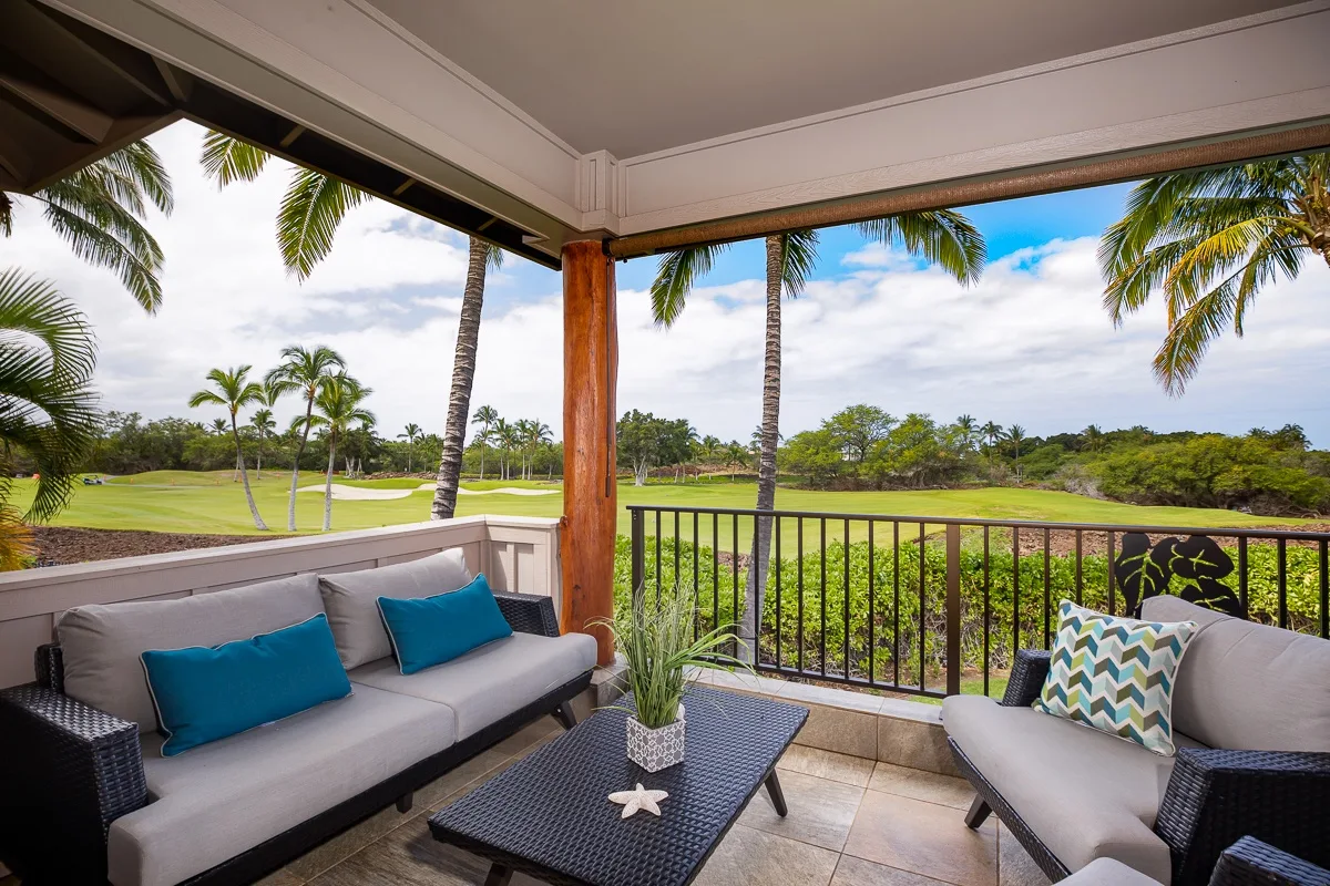 A covered patio has cushioned seating overlooking a green landscape and palm trees.