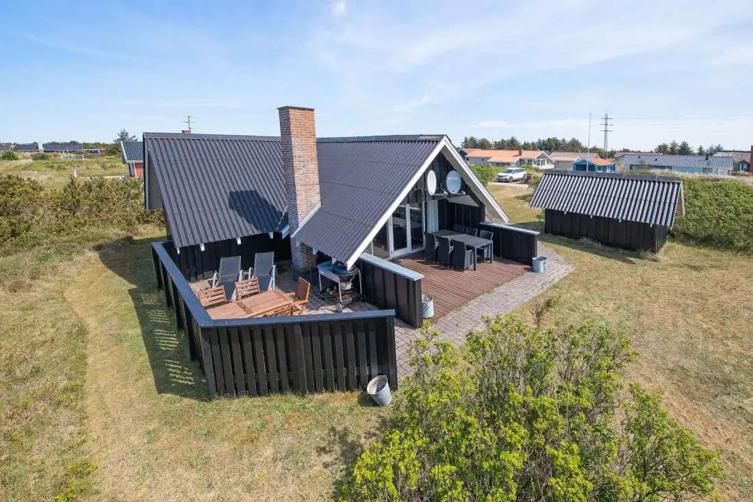 Una casa de techo oscuro con una terraza de madera se encuentra en una zona abierta con césped.