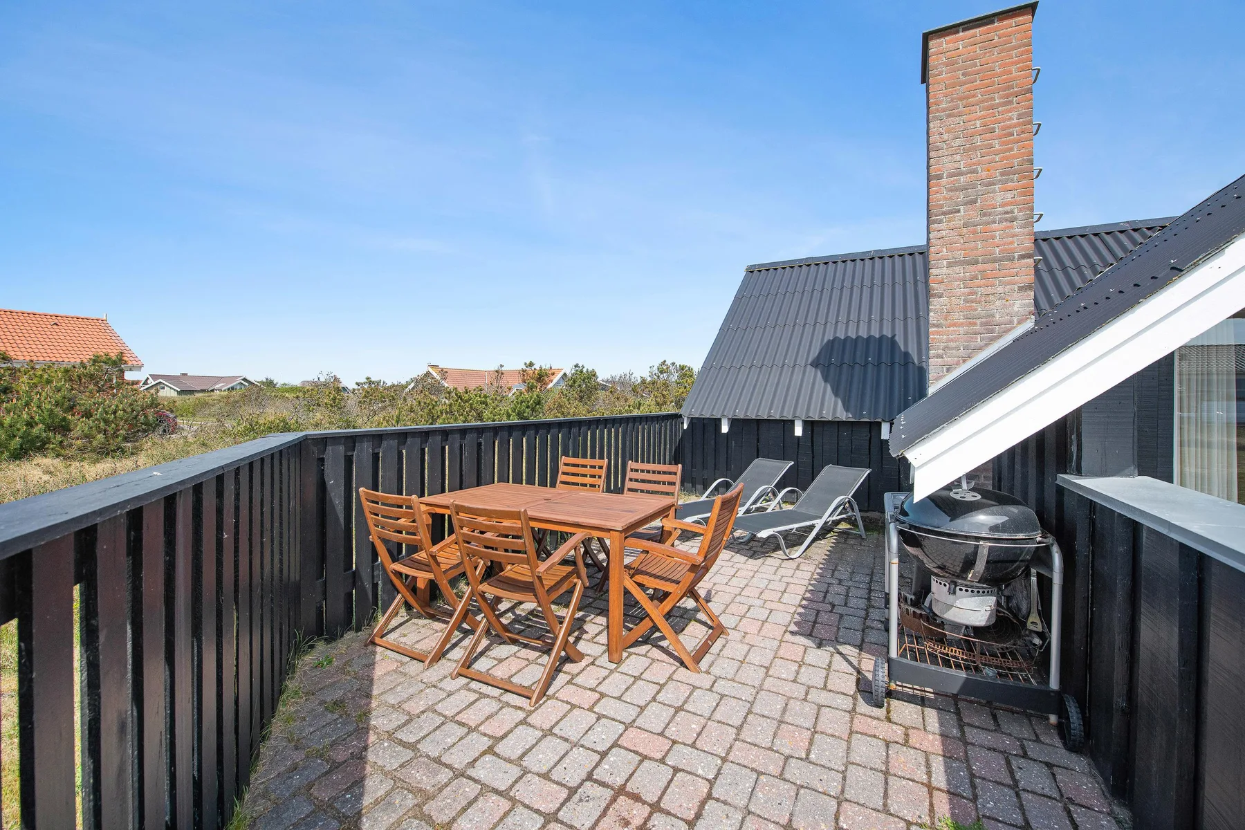 Una terraza pavimentada tiene asientos de exterior de madera y una parrilla junto a la casa.