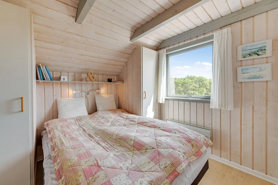 Un dormitorio con una cama bajo un techo inclinado y una ventana con vistas a los árboles.