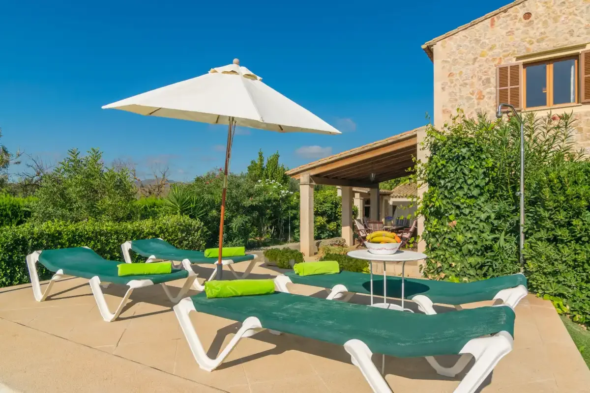 Un patio junto a la piscina con asientos reclinables, una sombrilla y vegetación.
