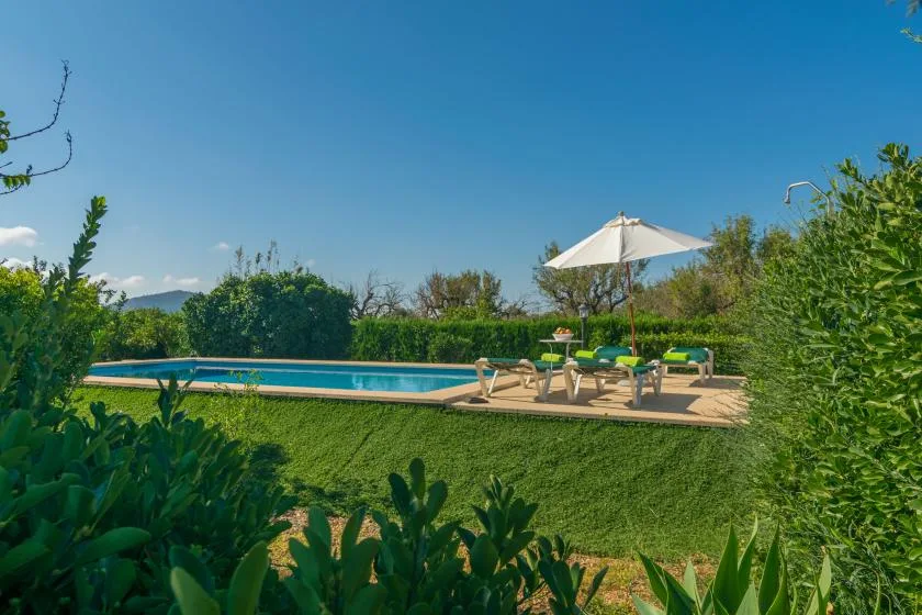 Una piscina al aire libre junto a un césped y una zona de descanso con sombra.