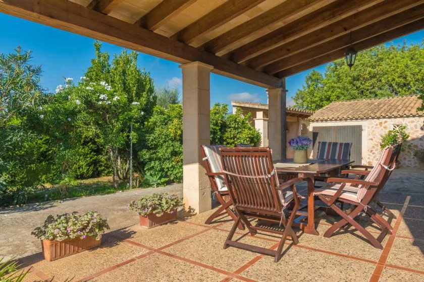 Un patio exterior cubierto con mesa y asientos de madera da a un jardín frondoso.