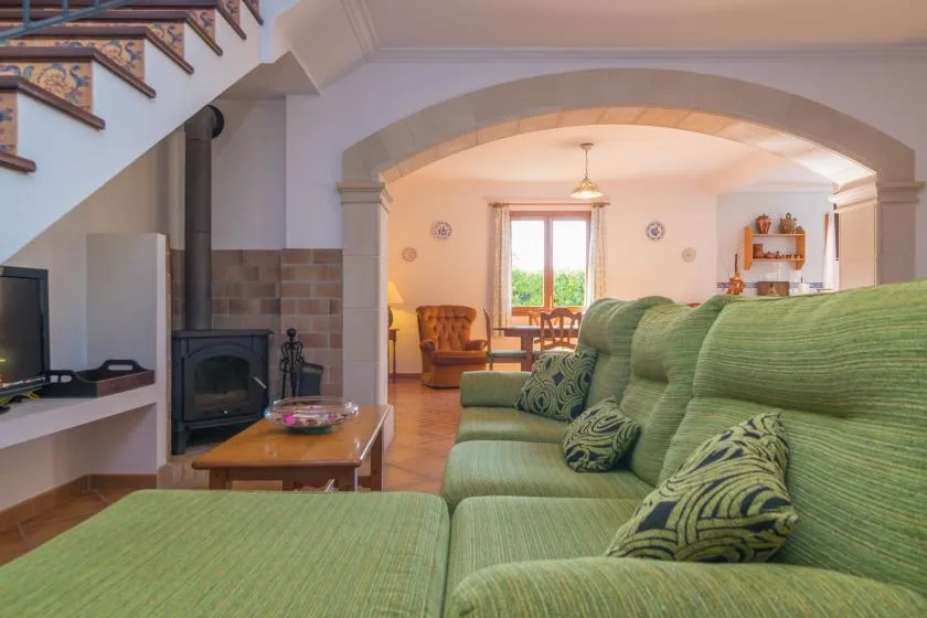 Una sala de estar con asientos verdes, una estufa de leña y un comedor al otro lado del arco.