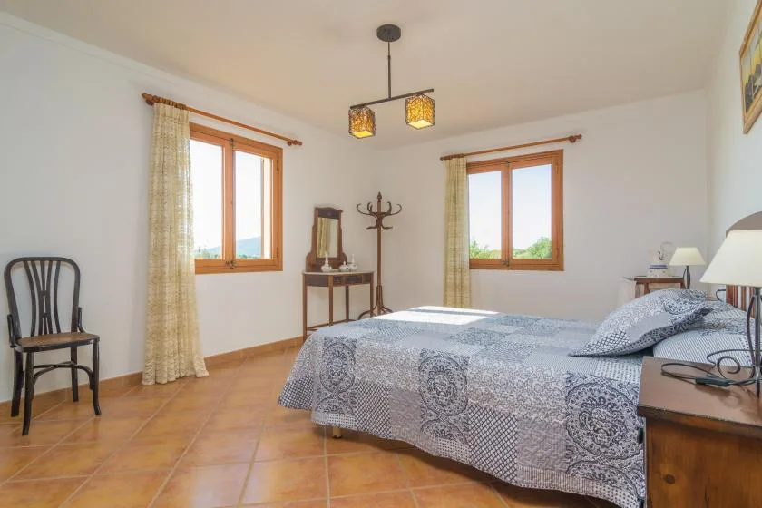Un dormitorio con una colcha estampada, dos ventanas y una silla de madera.