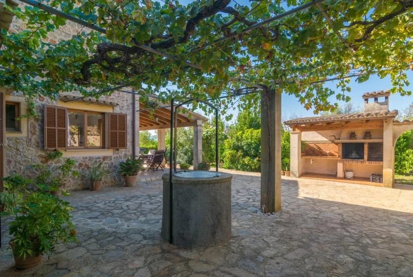 Un patio de piedra a la sombra de una pérgola frondosa, con un pozo y una zona de cocina exterior.