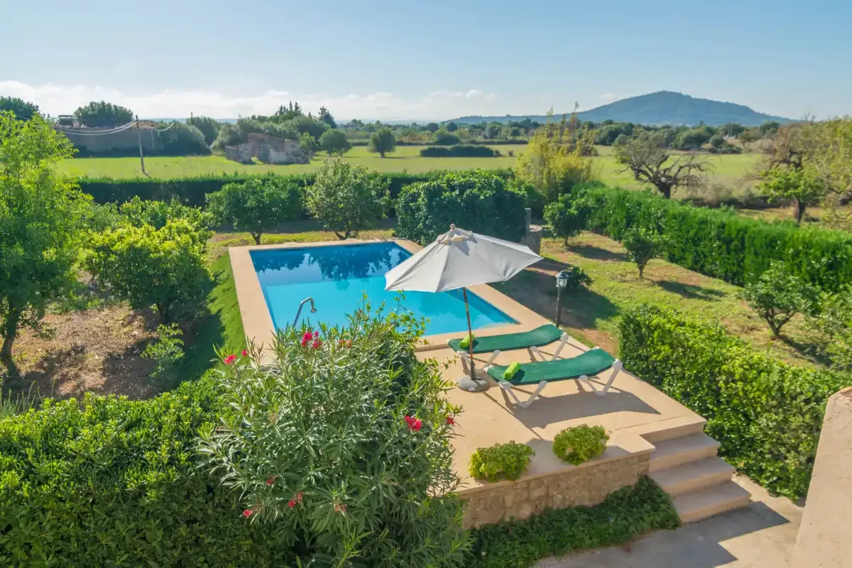 Una piscina con tumbonas y una sombrilla está rodeada de jardines verdes y campos.