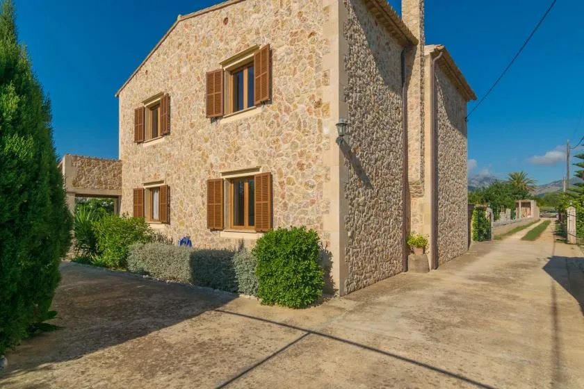 Exterior de una casa de piedra con contraventanas de madera junto a un camino pavimentado.
