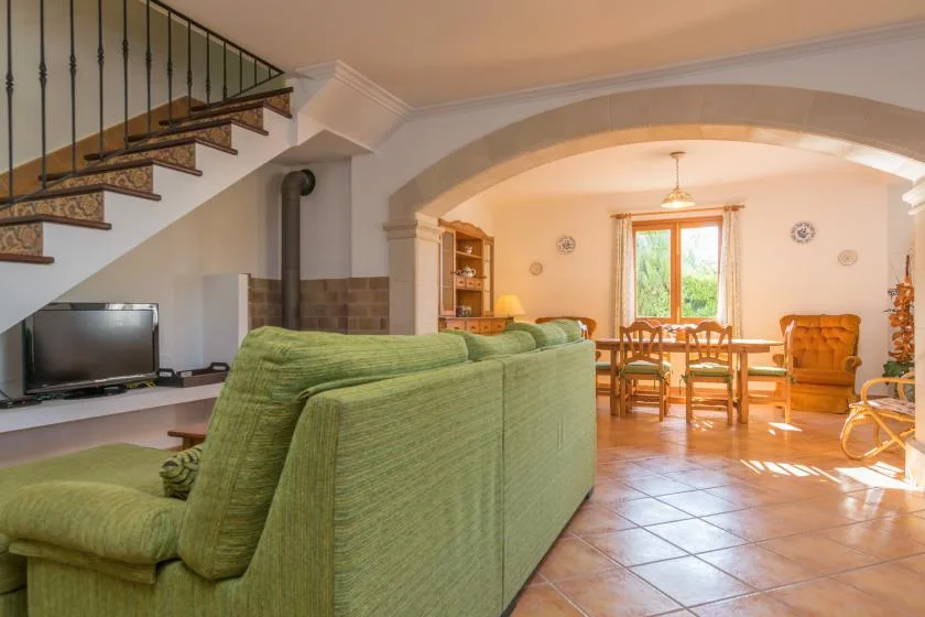 Una sala de estar con asientos verdes, un televisor y un comedor al otro lado de un arco.