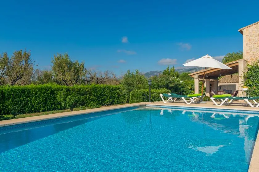 Una piscina exterior junto a un patio con tumbonas y una sombrilla.