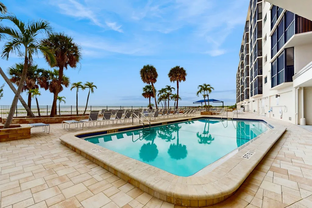 An outdoor swimming pool sits beside a paved deck and a palm-lined waterfront.