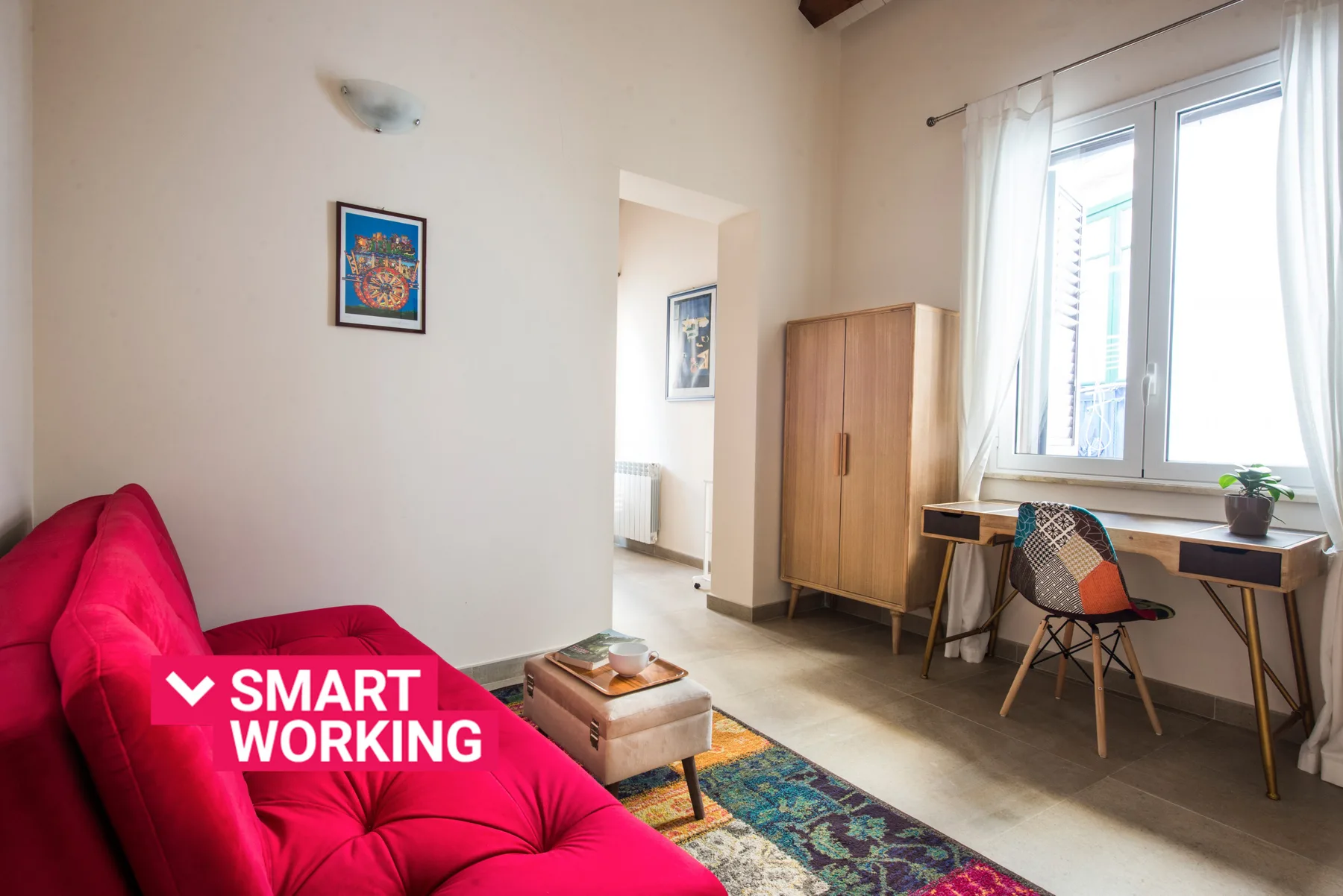 Una habitación luminosa tiene un sofá rojo y un escritorio junto a una ventana grande.